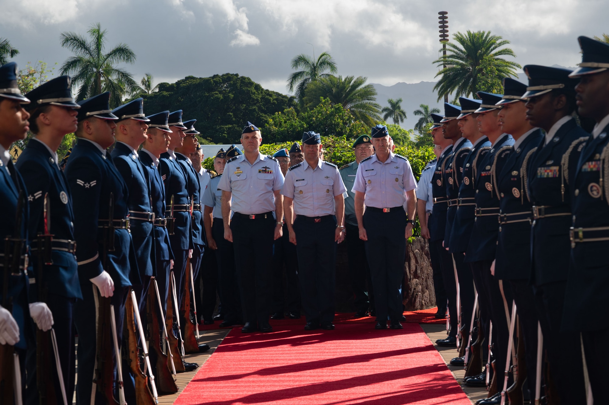 U.S. Air Force Chief of Staff Gen. Ken Wilsbach, U.S. Air Force Gen. Kevin Schneider, Pacific Air Forces commander, and Royal Australian Air Force Air Marshal Stephen Chappell, Chief of Air Force, stand together in uniform at the end of a red carpet. They are framed by two rows of an honor cordon made up of Airmen in dress uniforms, with a tropical background of palm trees and mountains.