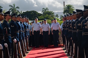 U.S. Air Force Chief of Staff Gen. Ken Wilsbach, U.S. Air Force Gen. Kevin Schneider, Pacific Air Forces commander, and Royal Australian Air Force Air Marshal Stephen Chappell, Chief of Air Force, stand together in uniform at the end of a red carpet. They are framed by two rows of an honor cordon made up of Airmen in dress uniforms, with a tropical background of palm trees and mountains.