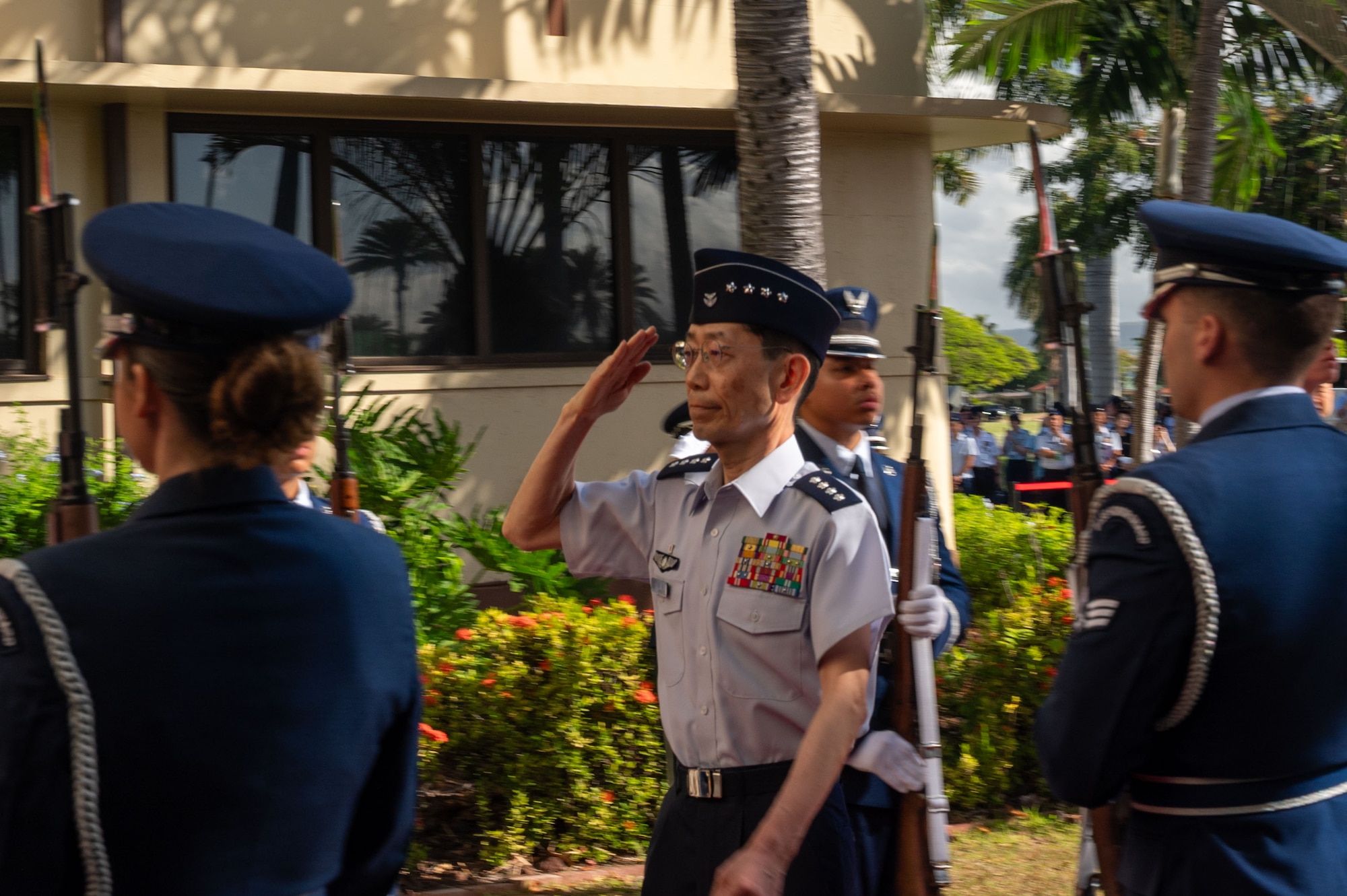 Japanese Air Self-Defense Force Gen. Takehiro Morita, in uniform, renders a salute as he walks past an honor cordon of U.S. Airmen. He is outdoors in a tropical setting with palm trees in the background.