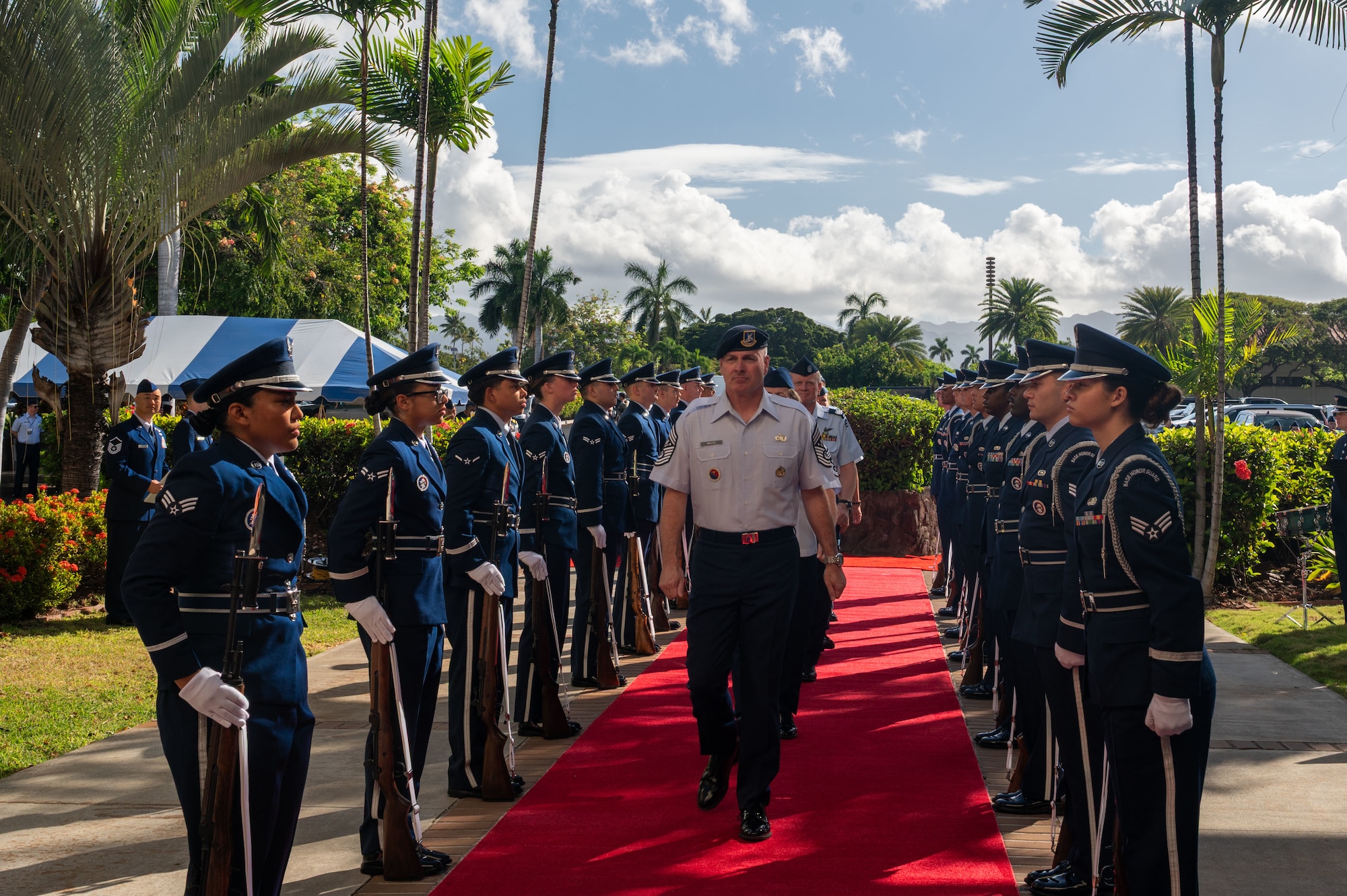 U.S. Air Force Chief Master Sergeant of the Air Force David Wolfe leads a group of senior enlisted leaders down a red carpet through an honor cordon. The cordon is formed by two rows of Airmen in dress uniforms holding rifles in an outdoor, tropical setting.