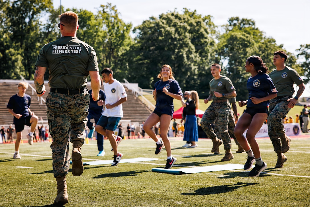 U.S. Marine Corps Staff Sgt. William Kearly, a force fitness instructor trainer with Training Command, leads warm-up exercises with students from Marine Corps Base Quantico’s Department of War Education Activity schools prior to conducting the Presidential Fitness Test at Butler Stadium on MCB Quantico, Virginia, May 12, 2026. The Presidential Fitness Test consists of three fitness components that are designed to strengthen health, physical fitness, resilience, and establish lifelong healthy habits. The PFT also recognizes multiple levels of achievements, including the Presidential Fitness Award, National Fitness Award, Completion recognition and improvement.  (U.S. Marine Corps photo by Cpl. Braydon Rogers)