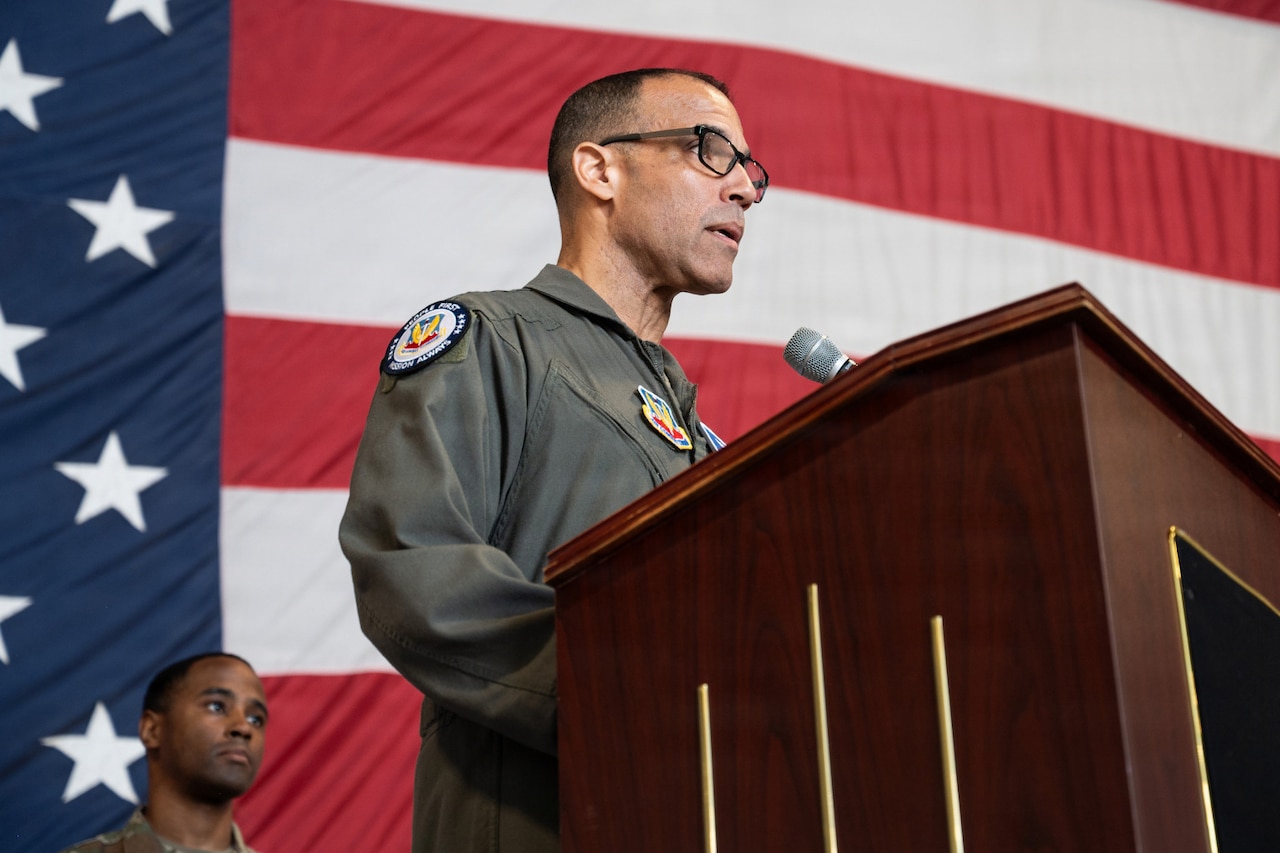 A man wearing a flight suit speaks into a microphone while standing behind a lectern; there is a large American flag in the background.