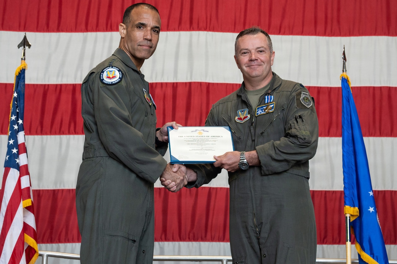 Two men wearing flight suits pose for a photo in front of a large American flag while holding an award.