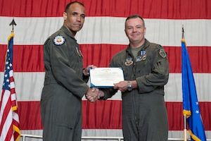 Two men wearing flight suits pose for a photo in front of a large American flag while holding an award.