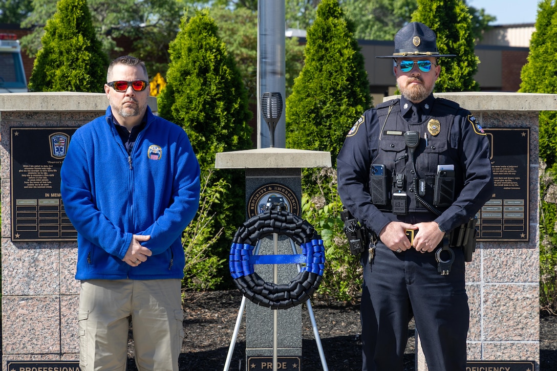 Two men stand with black and blue wreath between them. Man on left wearing sunglasses and blue fleece. Man on right wearing sunglasses and police uniform.
