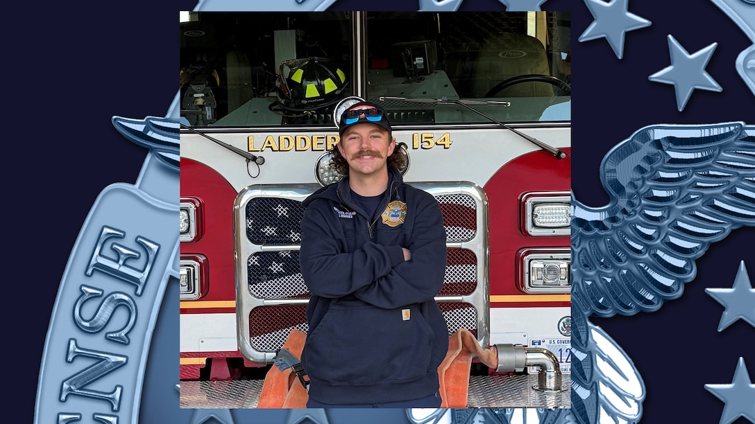 Man with mustache wearing ball cap and blue fire uniform stands in front of fire truck with arms crossed