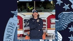 Man with mustache wearing ball cap and blue fire uniform stands in front of fire truck with arms crossed