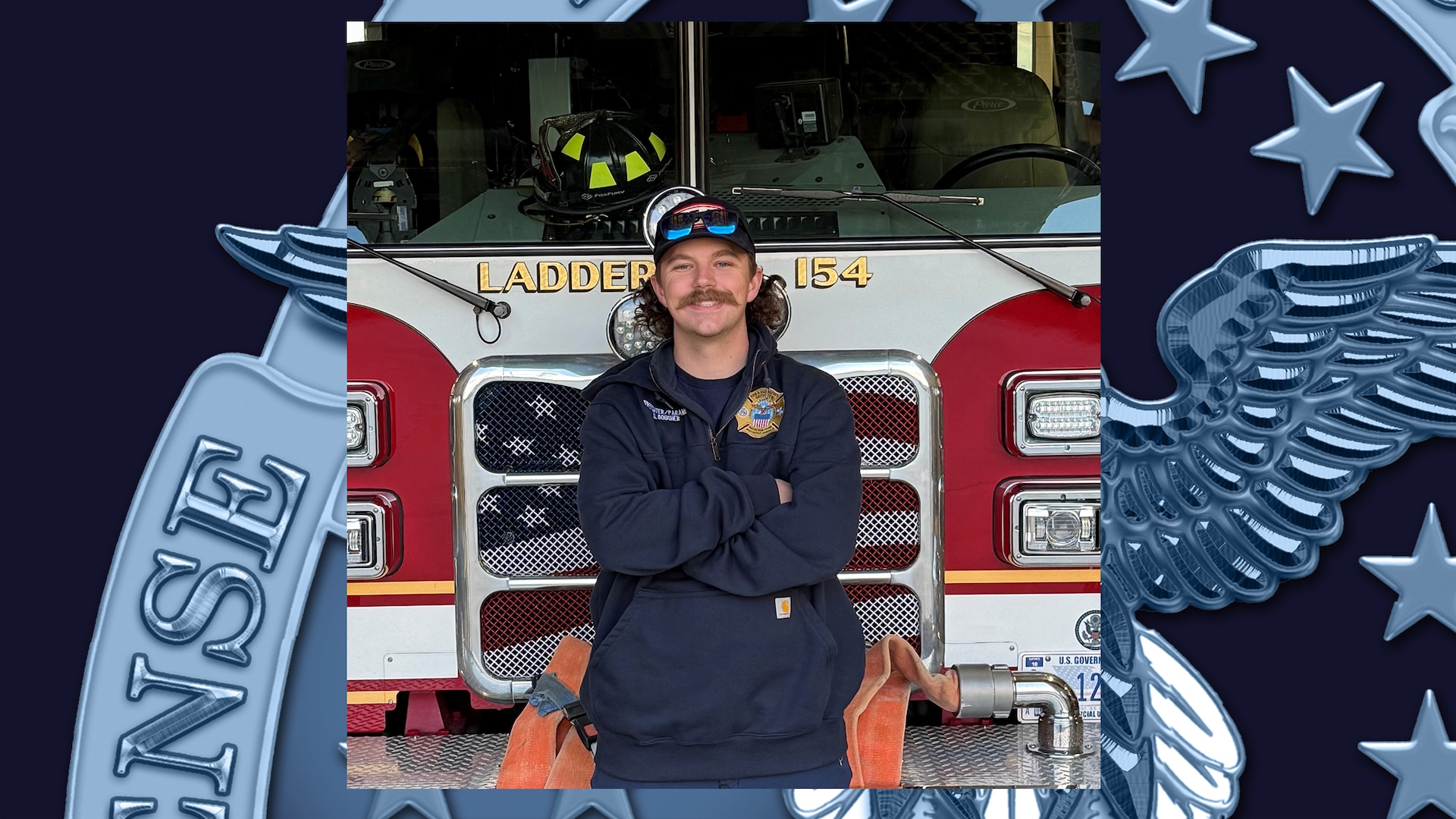 Man with mustache wearing ball cap and blue fire uniform stands in front of fire truck with arms crossed