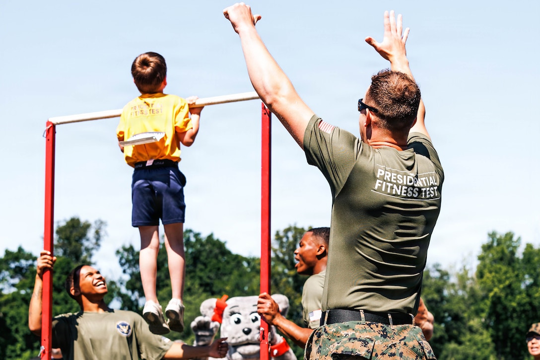 Three Marines cheer as a military child does a pullup against a blue sky.