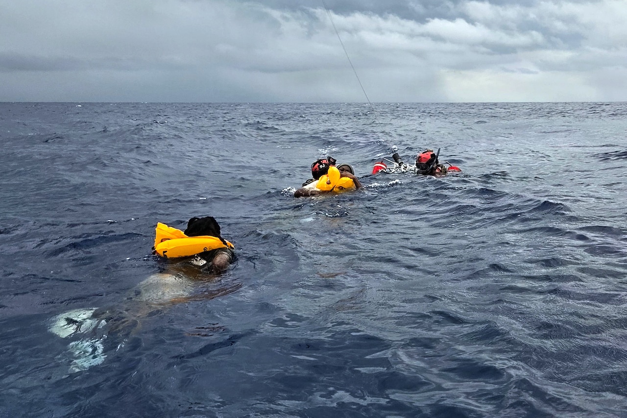 Two people in wet suits assist two other people with life preservers swimming in ocean into a recovery basket as a rescue helicopter hovers above.