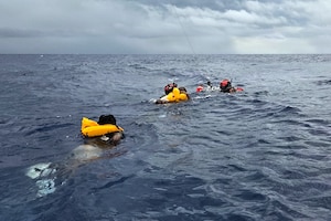 Two people in wet suits assist two other people with life preservers swimming in ocean into a recovery basket as a rescue helicopter hovers above.