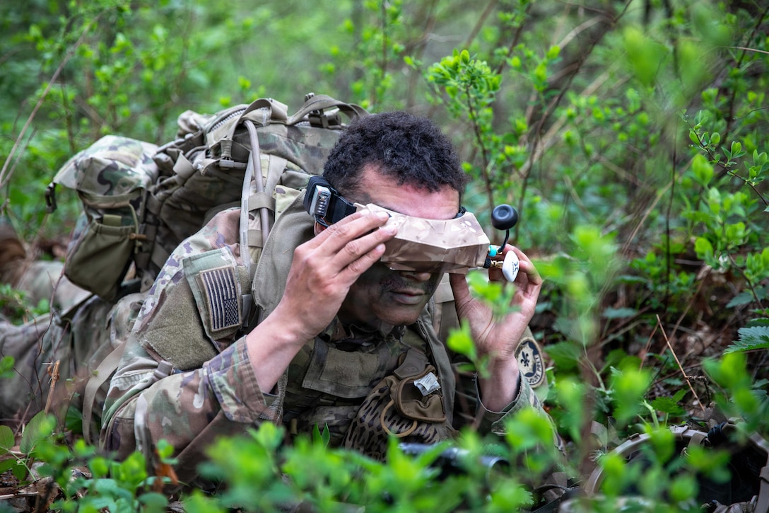 A soldier wearing tech goggles lies in a wooded area surrounded by greenery.