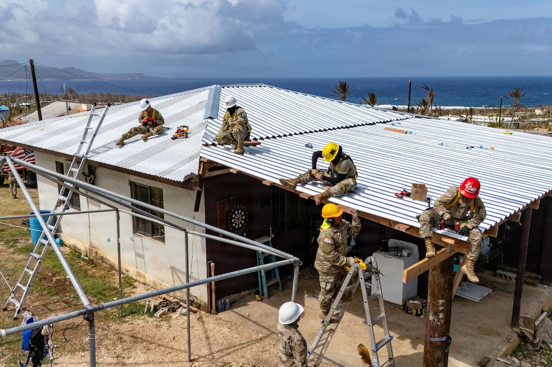 Soldiers in hard hats add a temporary roof to a home during the day with blue water and mountains in the background.