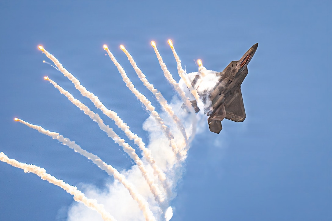 An aircraft performing a maneuver fires flares, leaving streaks in a blue sky.