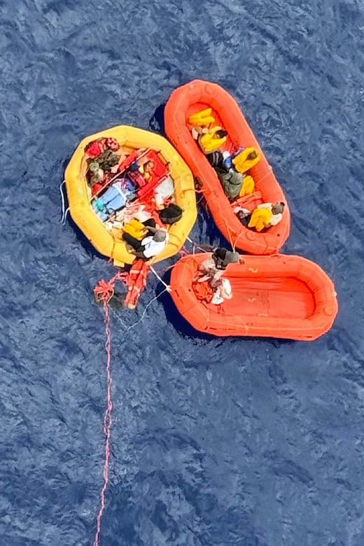 Two orange and one yellow rescue boats containing a group of people float in a large body of water.