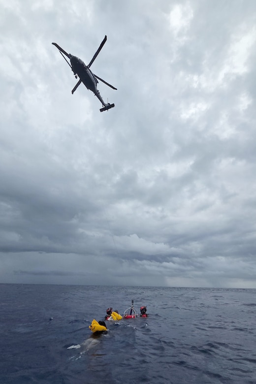 Two people in wet suits assist two other people with life preservers swimming in ocean into a recovery basket as a rescue helicopter hovers above.