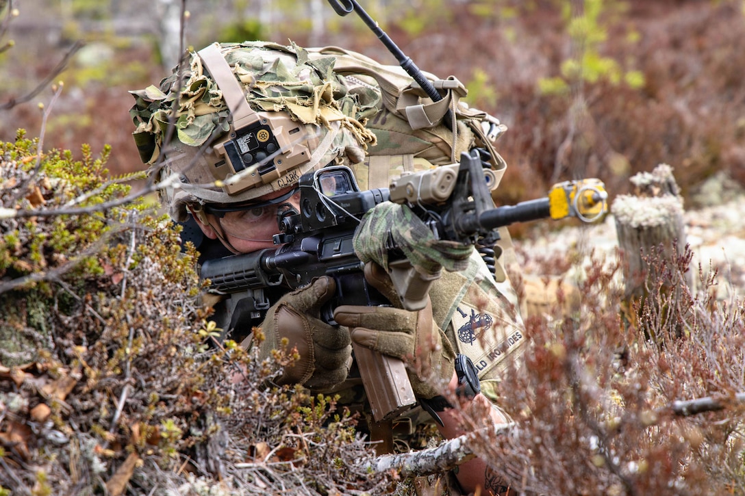 A soldier points a machine gun with a blank-firing adapter on it. The soldier’s face, hands and weapon are visible, with brush all around.