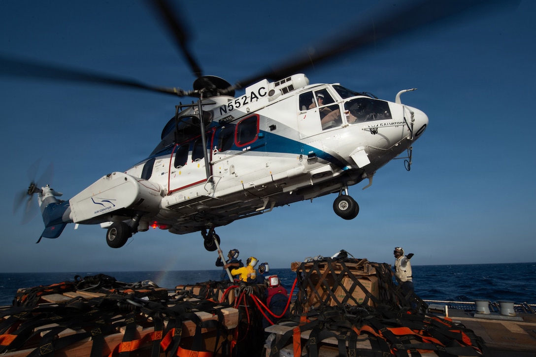 A helicopter hovers over a ship’s deck. People on the deck are surrounded by cargo, and one person uses a tool to hook onto the helicopter.