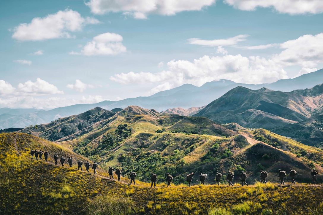 Twenty-some soldiers walk in a line on a path in the mountains, with green mountain peaks in the background.