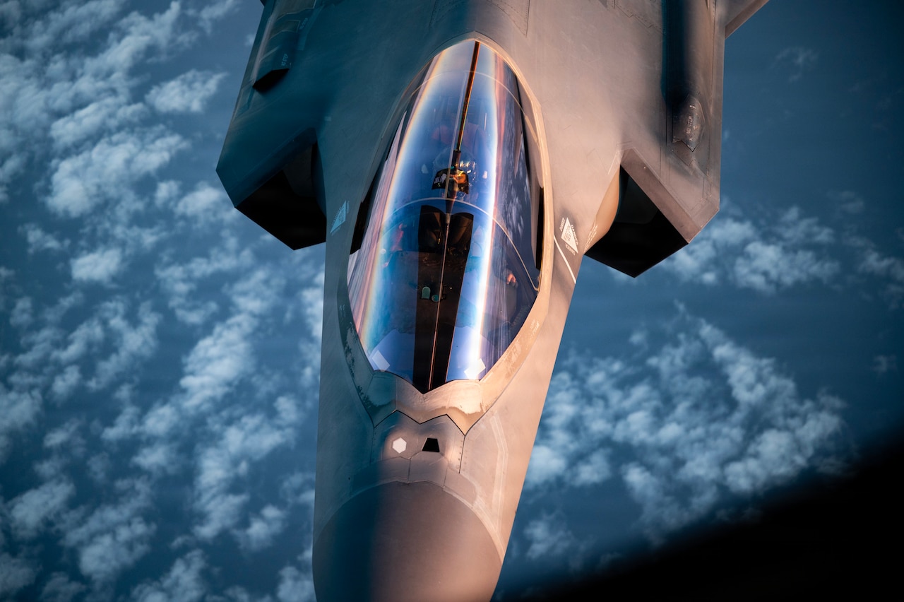 A person in a flight suit and helmet gazes up through the cockpit of a fighter jet.