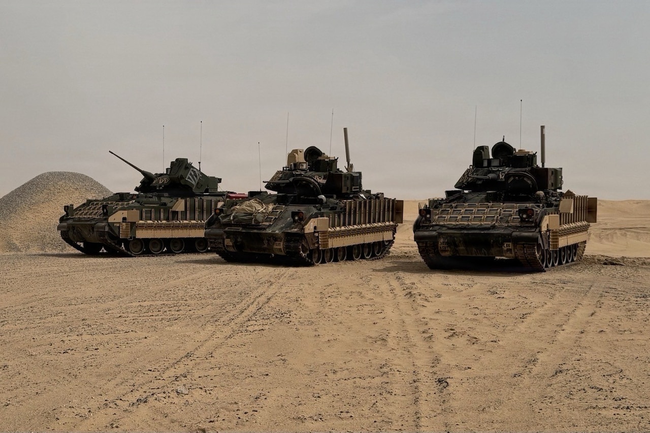 Three armored combat vehicles sit near each other in a desert environment.