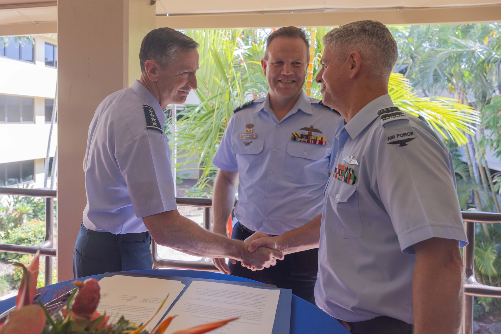 U.S. Pacific Air Forces commander, Gen. Kevin Schneider, and Royal New Zealand Air Force Air Vice-Marshal Darryn Webb shake hands with smiles after signing documents, while Royal Australian Air Force Air Marshal Stephen Chappell looks on smiling. The signed papers are visible on the table in front of them, with a tropical background.