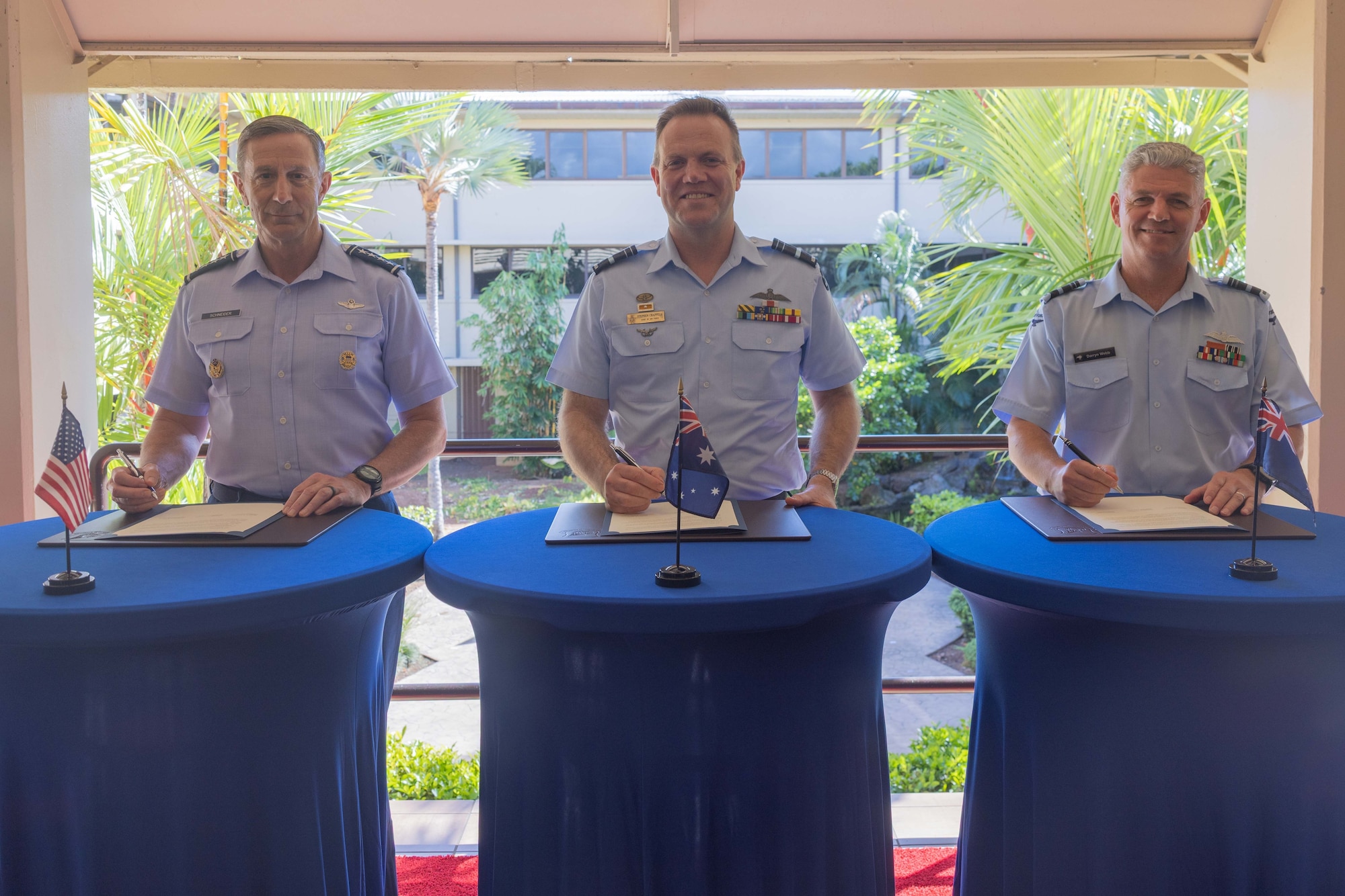 U.S. Pacific Air Forces commander, Gen. Kevin Schneider; Royal Australian Air Force Air Marshal Stephen Chappell; and Royal New Zealand Air Force Air Vice-Marshal Darryn Webb stand behind individual tables with documents prepared for signing. Each table displays the national flag corresponding to the leader's respective country, set against a background of lush tropical foliage.