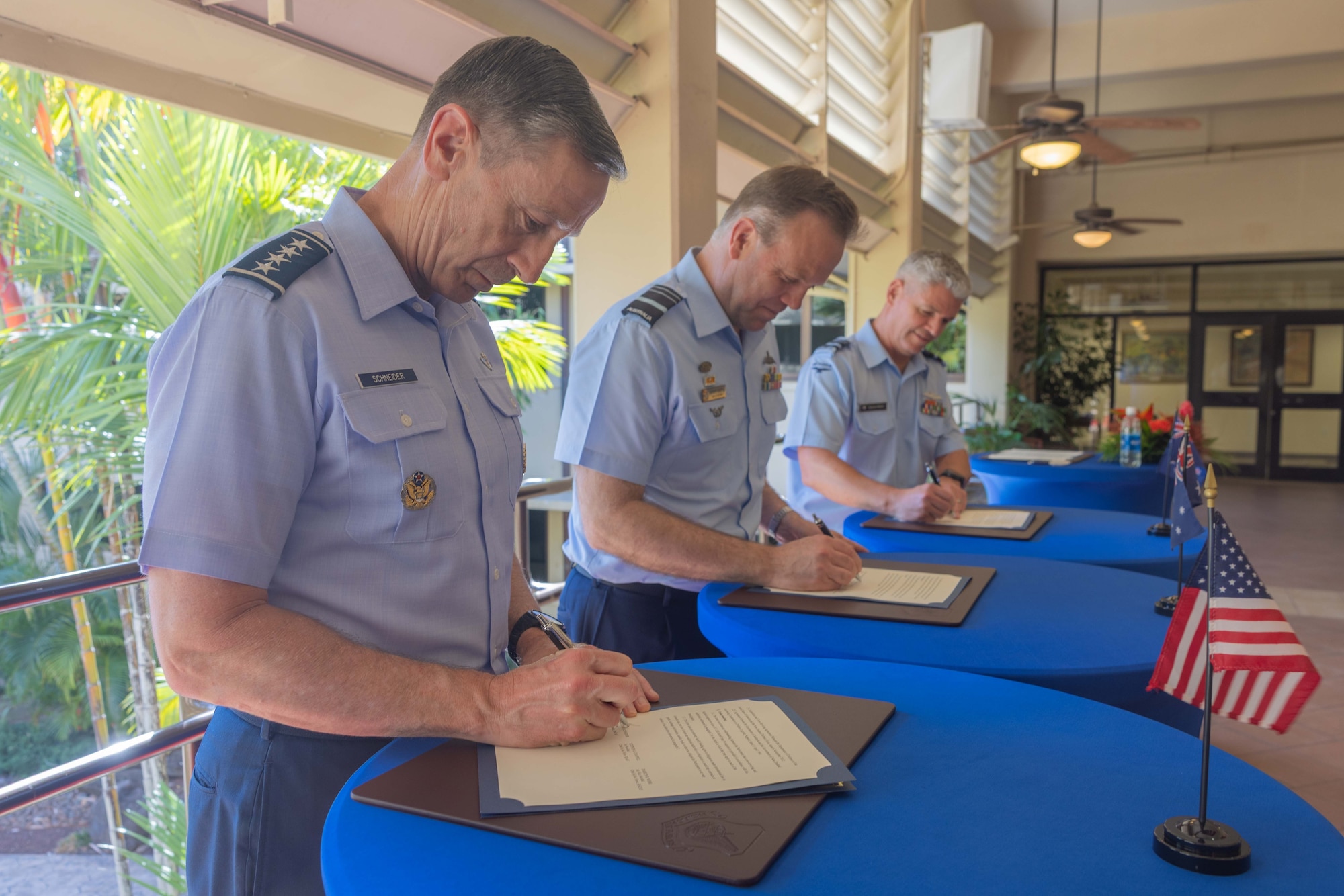 U.S. Pacific Air Forces commander, Gen. Kevin Schneider; Royal Australian Air Force Air Marshal Stephen Chappell; and Royal New Zealand Air Force Air Vice-Marshal Darryn Webb stand in a line, simultaneously signing documents at individual tables. Small flags of the United States, Australia, and New Zealand are visible on the tables, with a tropical background.