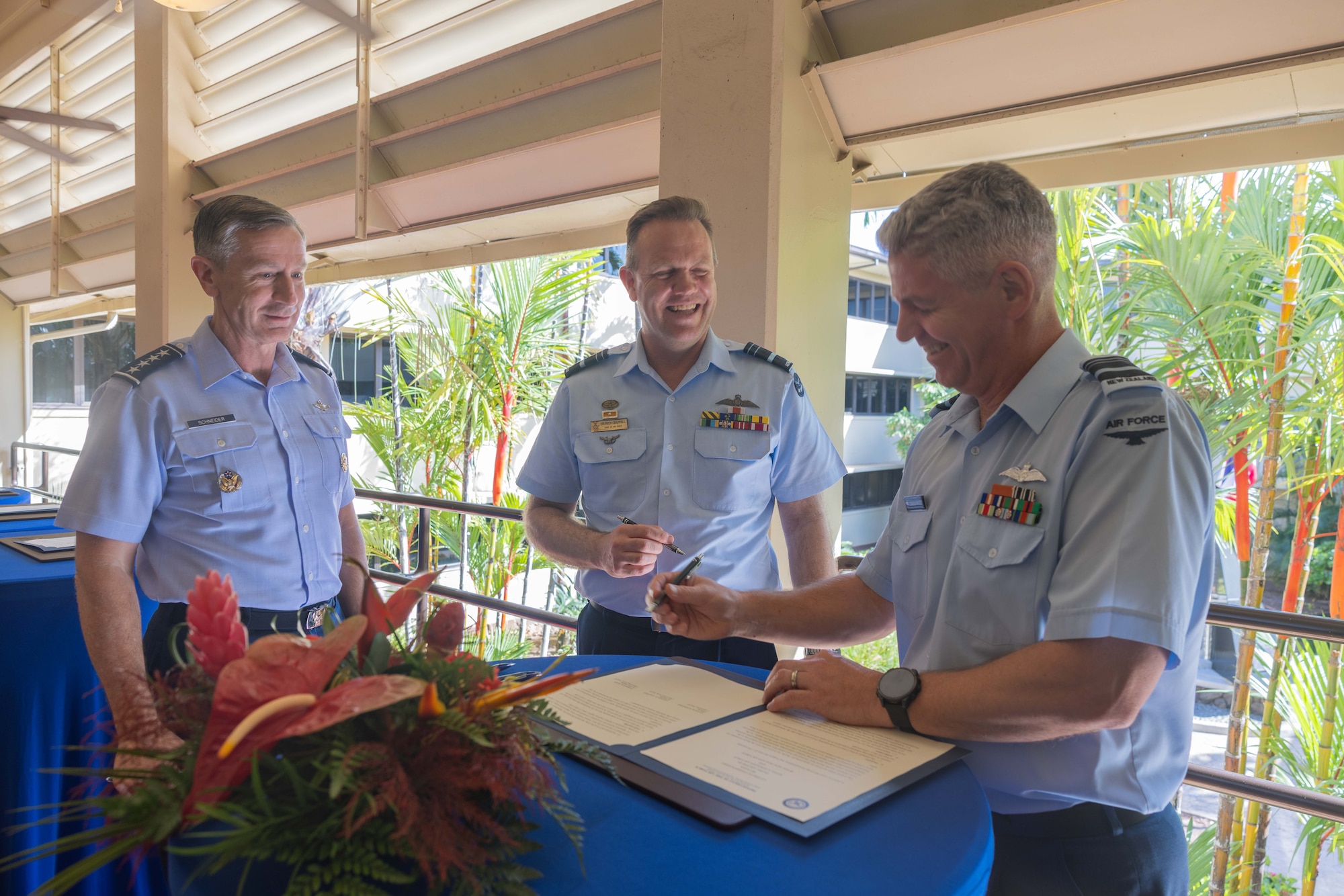 Royal New Zealand Air Force Air Vice-Marshal Darryn Webb smiles as he signs a document on a table, while Royal Australian Air Force Air Marshal Stephen Chappell and U.S. Pacific Air Forces commander, Gen. Kevin Schneider, look on. The three leaders are in uniform and gathered in a tropical setting.