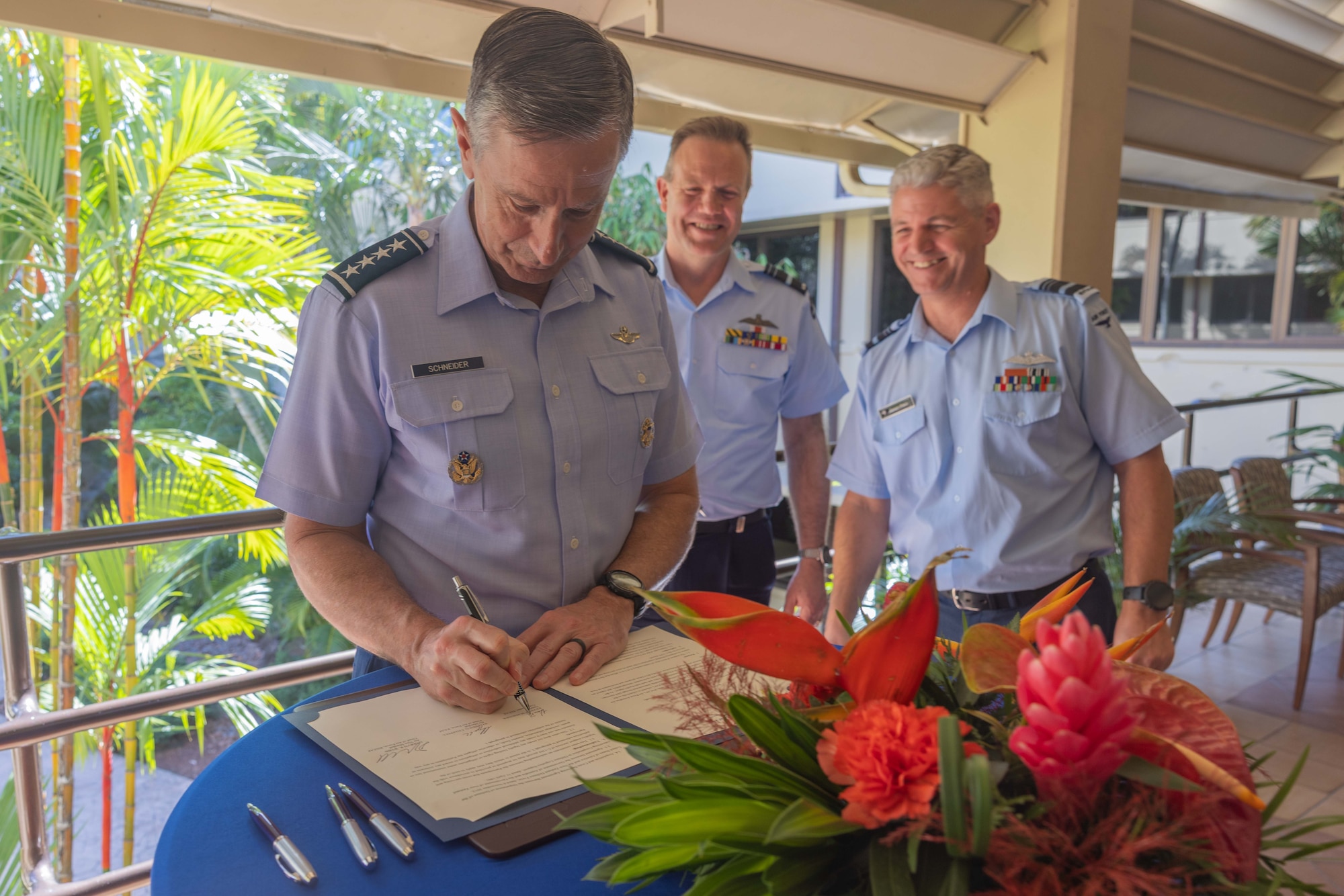 U.S. Pacific Air Forces commander, Gen. Kevin Schneider, signs a document on a table adorned with a large tropical flower arrangement. Royal Australian Air Force Air Marshal Stephen Chappell and Royal New Zealand Air Force Air Vice-Marshal Darryn Webb stand nearby, smiling. The three leaders are in uniform against a background of lush palm trees.