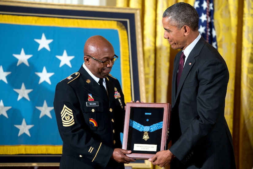 Two men hold a framed military medal with a large, framed flag behind them.