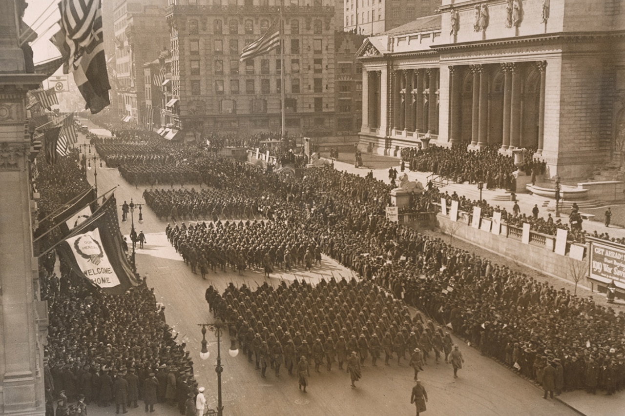 Soldiers march in large formations on a big city street as crowds look on.