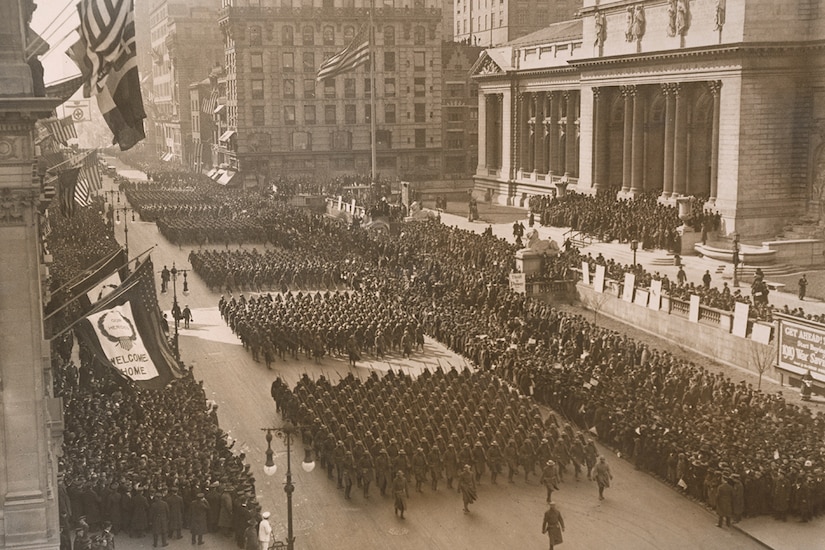 Soldiers march in large formations on a big city street as crowds look on.