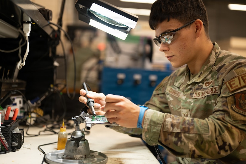 A service member wearing a camouflage military uniform uses a soldering tool on a circuit board. A magnifying light is above his hands.
