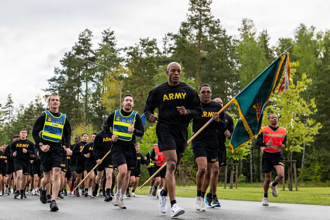 People in black T-shirts and shorts with Army imprinted in yellow on them run on a black path, with grass and tall trees in the background.