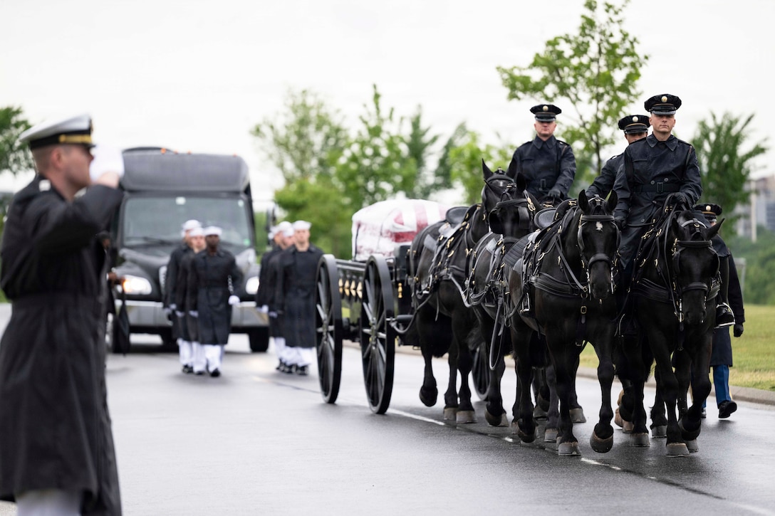 Service members operate a horse carriage with a casket wrapped in an American flag as fellow service members march in formation in front of a black vehicle.