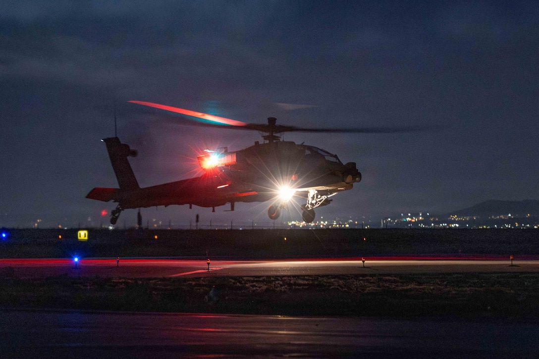 A helicopter illuminated by white and red lights hovers over a tarmac in a dark sky with city lights in the distance.