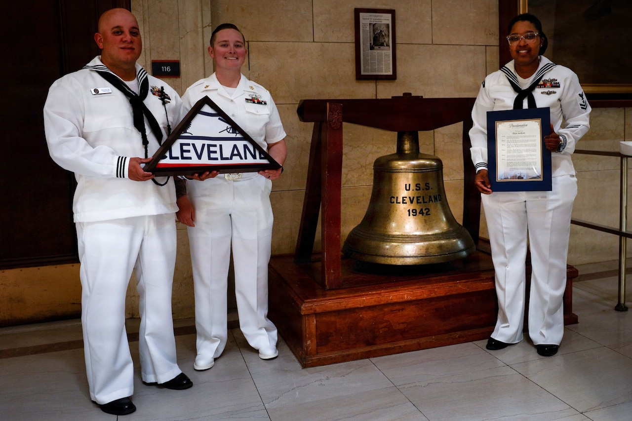 Three people wearing white Navy uniforms stand next to a large bell. Two sailors are holding a folded Cleveland flag and the other is holding a framed proclamation.