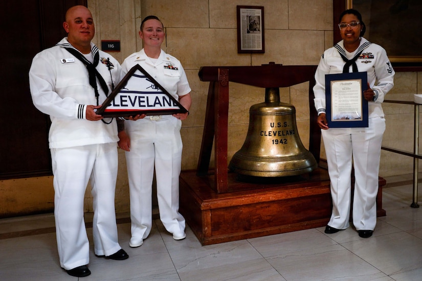 Three people wearing white Navy uniforms stand next to a large bell. Two sailors are holding a folded Cleveland flag and the other is holding a framed proclamation.