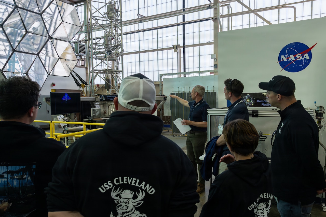 People wearing civilian attire watch as a man dressed in similar attire points and speaks during a tour of a facility.