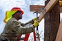 A Soldier in a red hard hat is seen standing on a ladder hammering in a nail for a support beam that is being used for a temporary roof.
