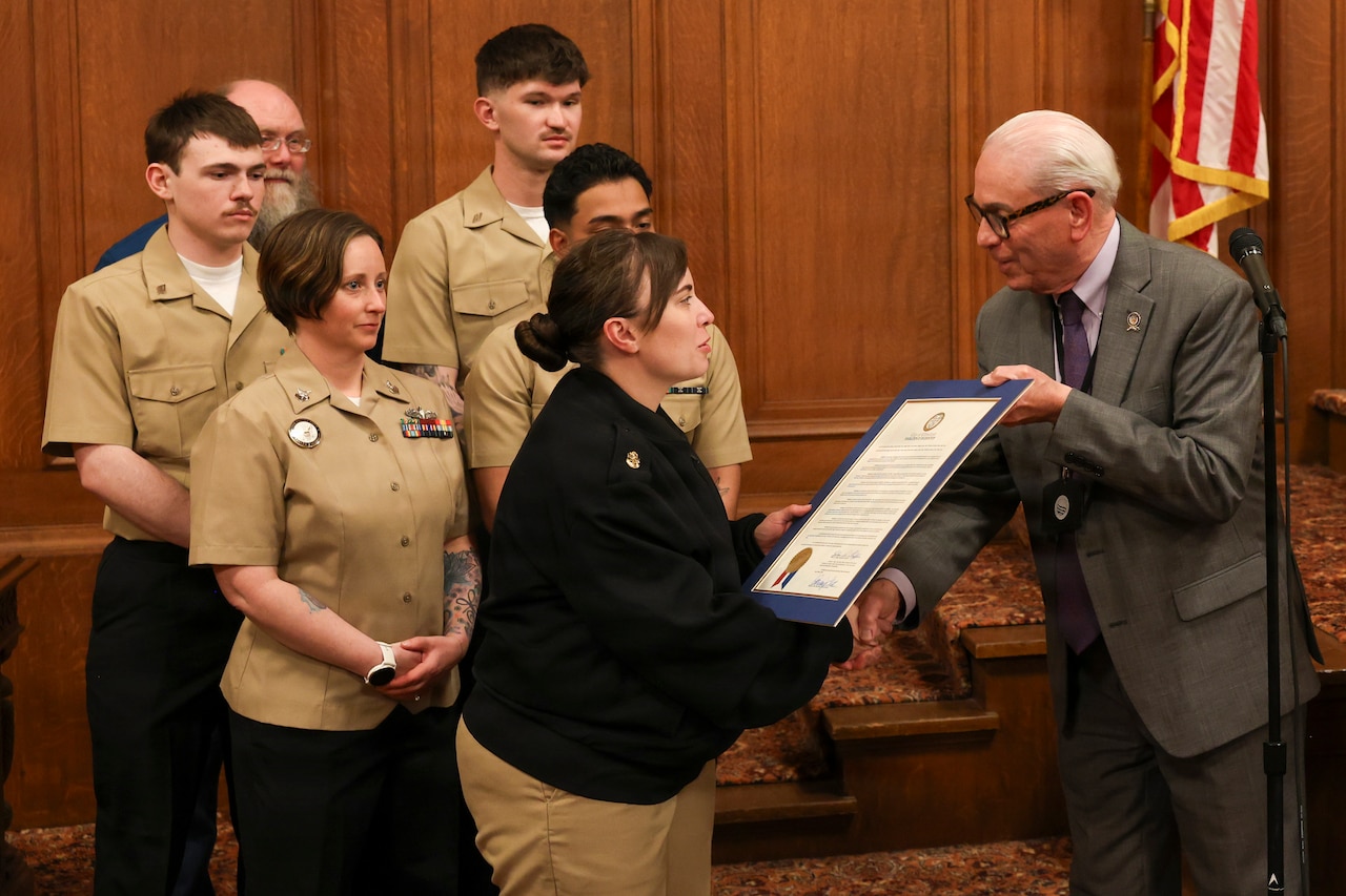 A man wearing a business suit presents a framed document to a woman wearing a khaki Navy uniform, while several other people also wearing similar Navy uniforms stand behind them, watching.