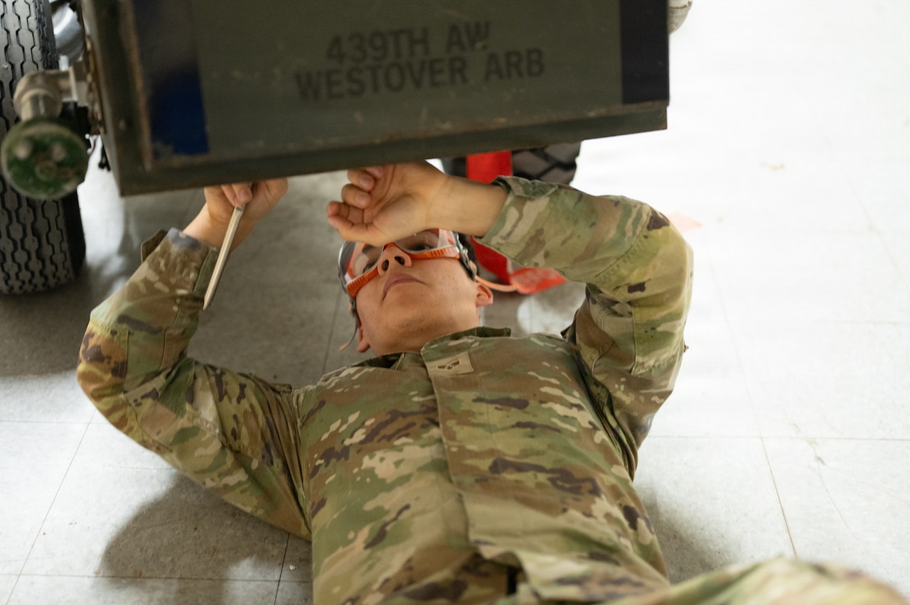 A service member in camouflage uniform wearing safety goggles lies on the floor under a cart.