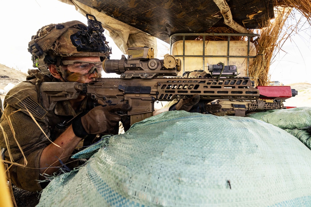 A soldier points a machine gun from behind a large sandbag or barrier, with another soldier and straw in the background.