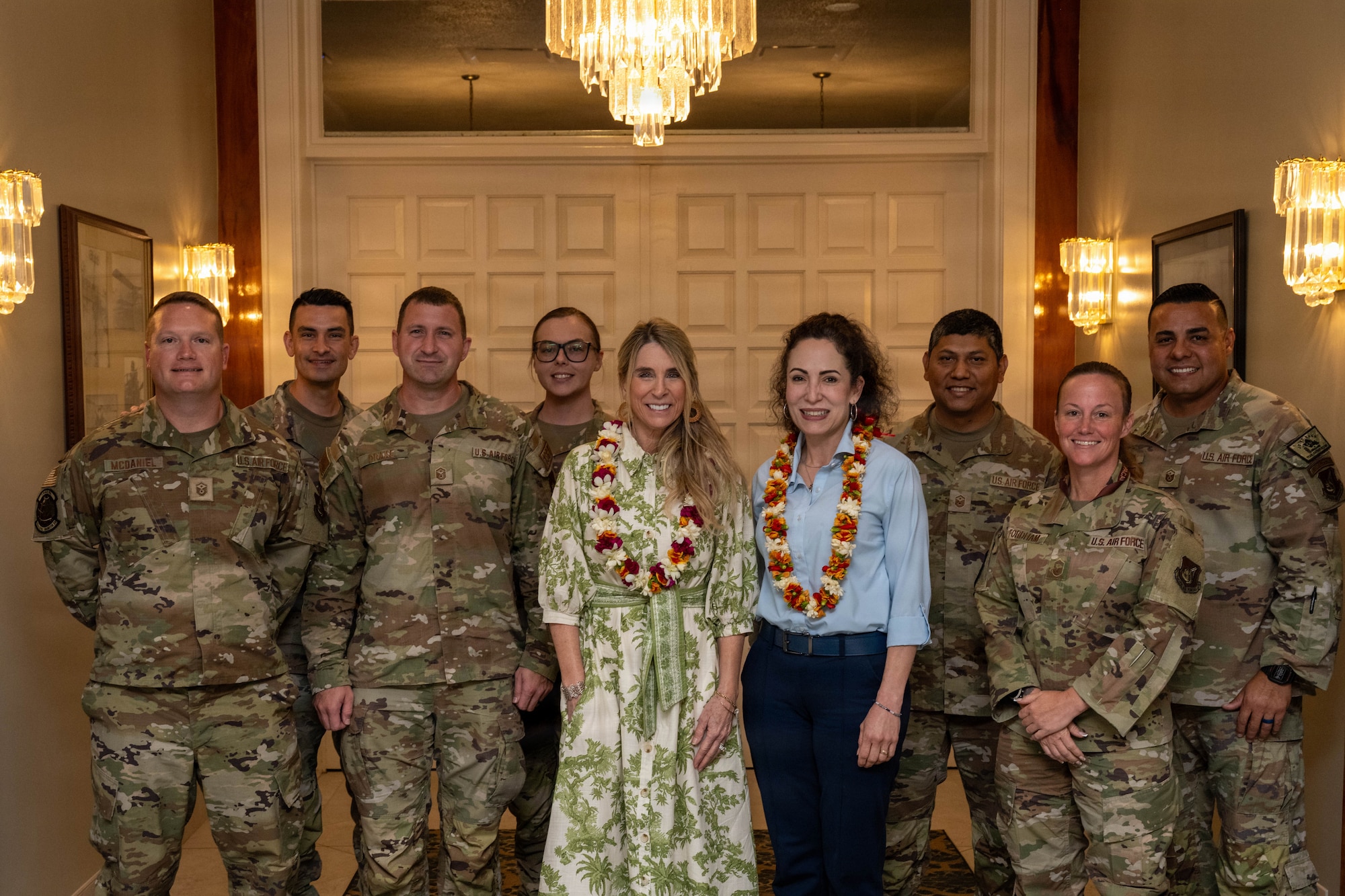 Two civilians stand with a group of military members for a group photo inside a building with warm lighting.