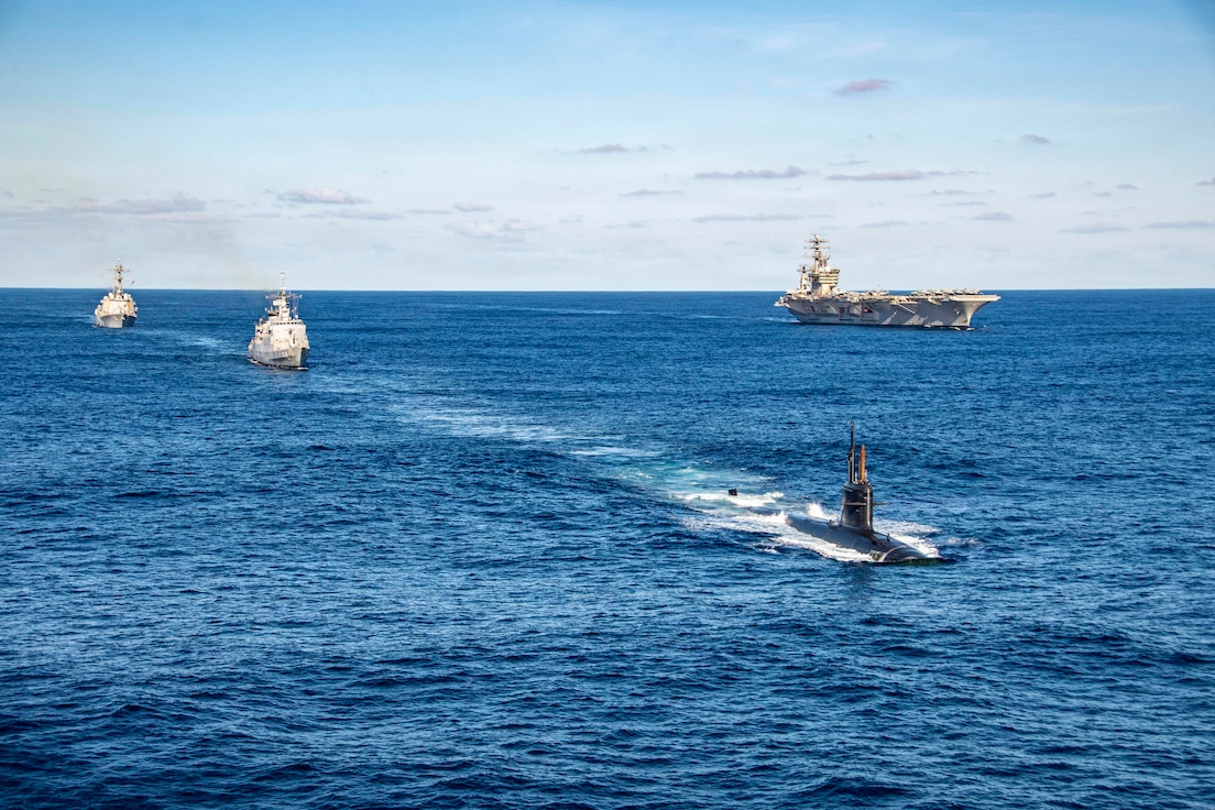 One large military ship steams on blue water alongside two smaller ships and a submarine, with the horizon in the background.