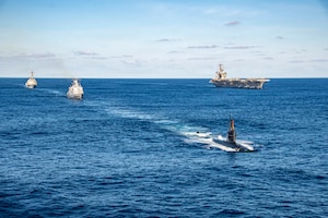 One large military ship steams on blue water alongside two smaller ships and a submarine, with the horizon in the background.