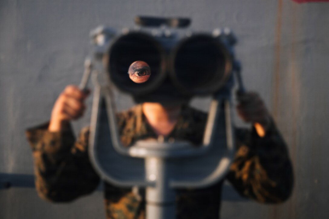 A Marine looks through ship-mounted binoculars, giving a magnified view of one eye.