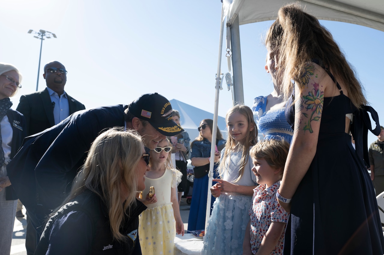 Secretary of War Pete Hegseth and Jennifer Hegseth smile while talking to a smiling child with an adult amid a crowd on a pier.