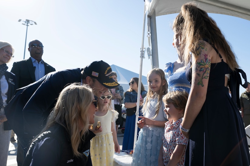Secretary of War Pete Hegseth and Jennifer Hegseth smile while talking to a smiling child with an adult amid a crowd on a pier.